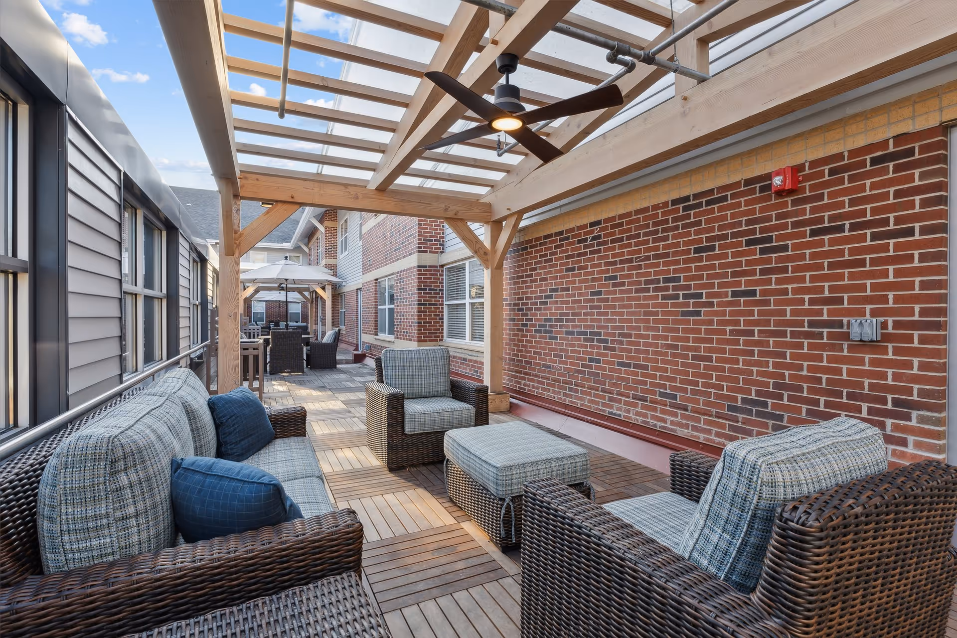 Outdoor patio area with wicker furniture including a sofa, armchairs, and an ottoman under a wooden pergola with a ceiling fan. The patio has wooden flooring tiles and is adjacent to a brick wall and a building with siding and windows. There are additional seating areas with umbrellas further along the patio.
