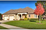 Single-story brick house with a three-car garage, a well-maintained green lawn, and a small tree with red leaves in the front yard under a clear blue sky.