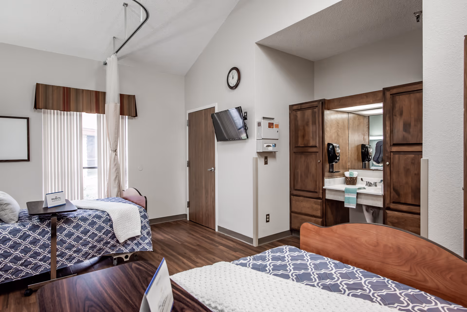 Interior nursing facility bedroom with two beds, overbed tables, a wall-mounted TV, and a vanity with wooden cabinets and sink.