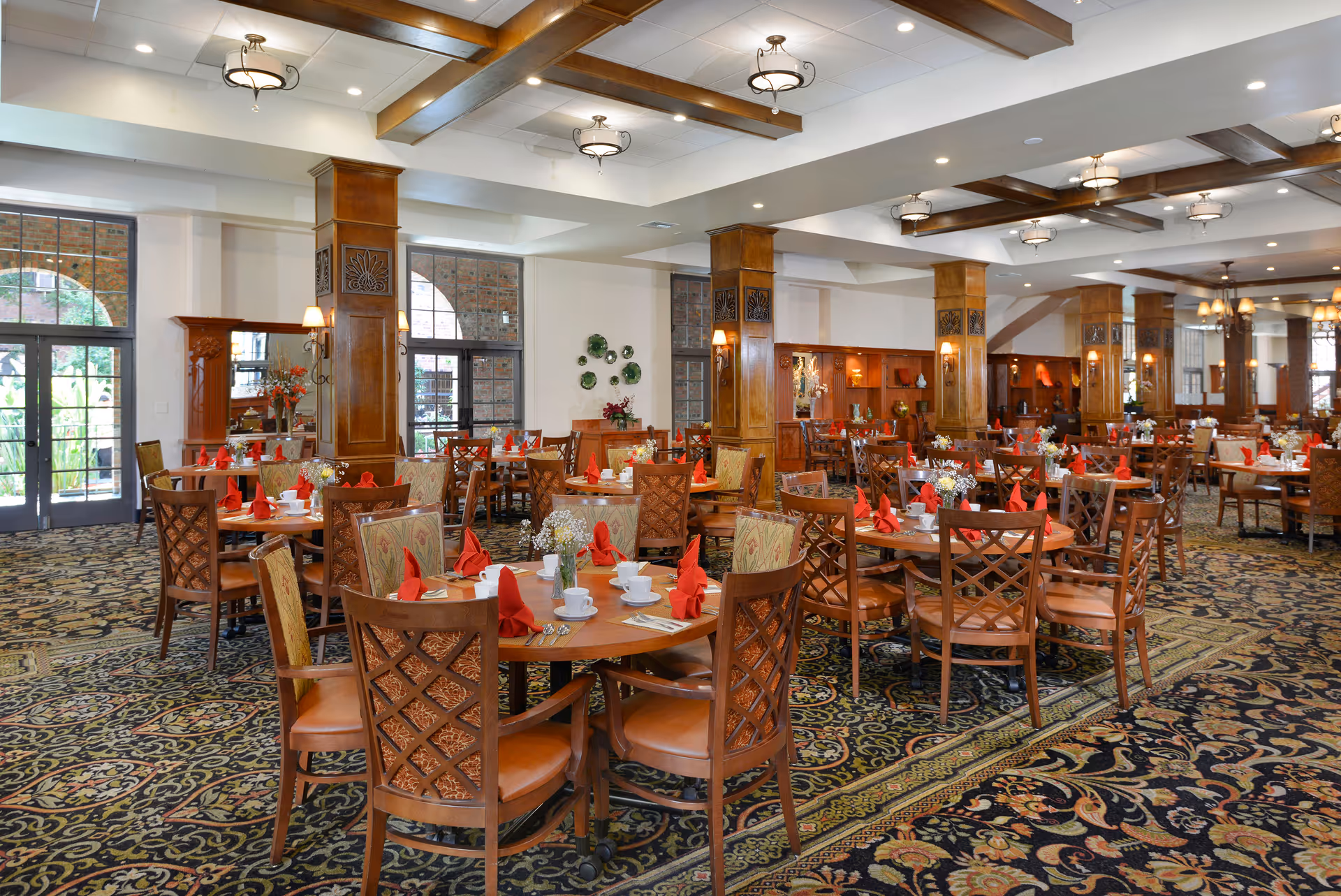 A spacious dining room with multiple round wooden tables set with white cups, plates, and red folded napkins. The room features patterned carpet flooring, wooden columns, large windows with arched tops, and ceiling lights with decorative fixtures. The atmosphere is elegant and inviting.