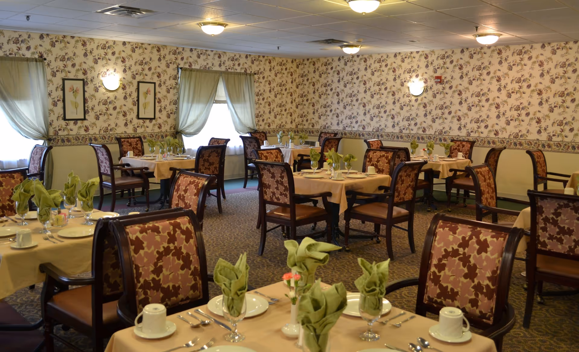 Dining room with multiple tables covered in beige tablecloths, each set with plates, cups, silverware, and green folded napkins. The room has floral wallpaper, patterned carpet, and windows with light green curtains. Chairs have a floral upholstery pattern.