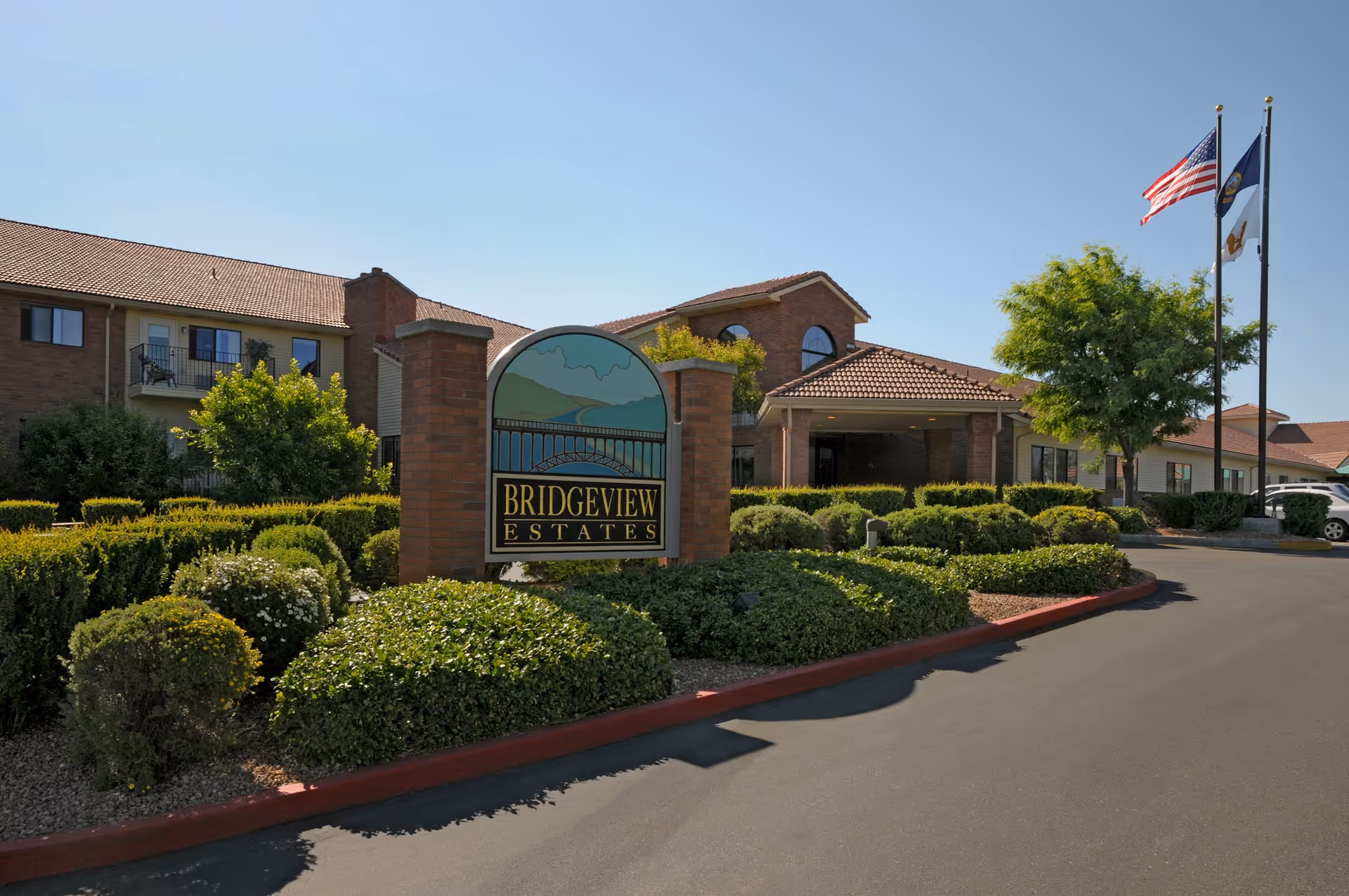 Exterior view of Bridgeview Estates senior living facility with a brick sign in the foreground surrounded by well-maintained bushes and greenery. The building has a tiled roof and multiple windows. Three flagpoles with the American flag and other flags are visible on the right side under a clear blue sky.