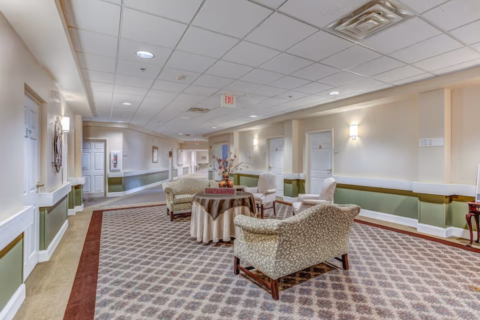 A well-lit hallway in a senior living facility with patterned carpet and beige walls featuring green wainscoting. There are several closed white doors along the hallway and a seating area with four upholstered chairs arranged around a round table covered with a tablecloth and a floral centerpiece. The ceiling has recessed lighting and an exit sign is visible in the distance.