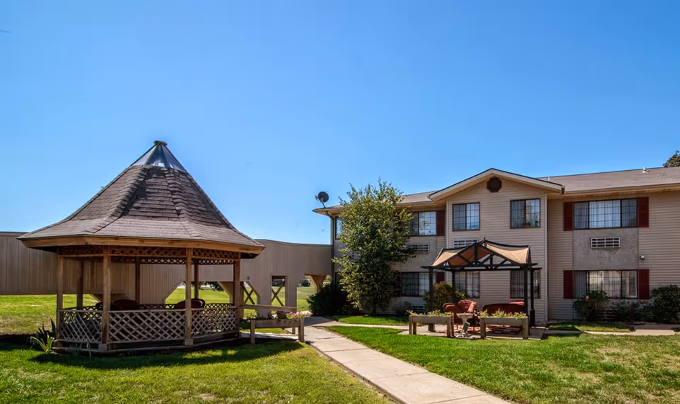 Outdoor courtyard with a wooden gazebo, seating area, walkway, and a two-story building under a clear blue sky.