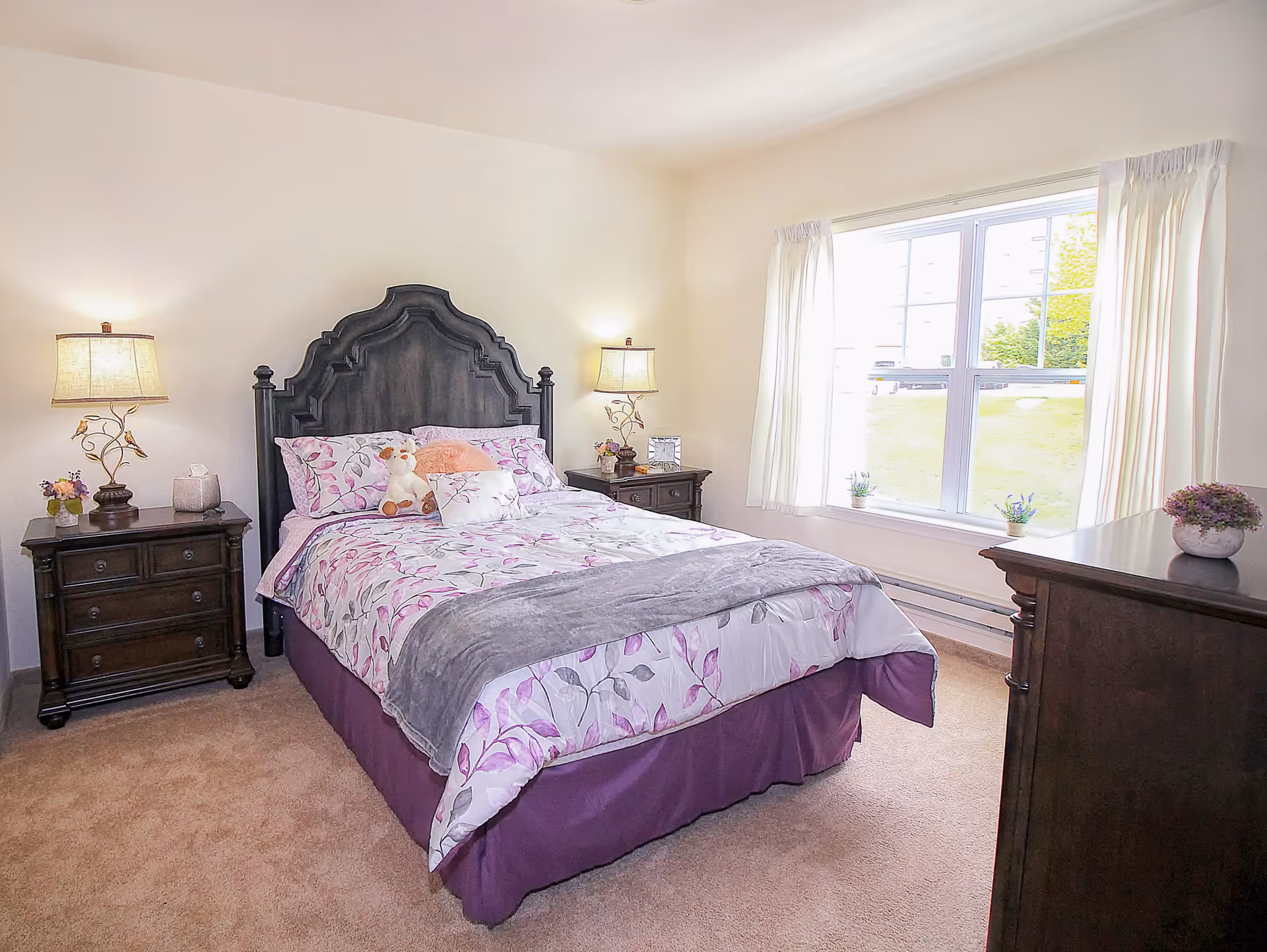 A bright bedroom with a large window letting in natural light. The room features a bed with a dark wooden headboard, floral bedding in shades of purple and pink, and a gray throw blanket. There are two matching dark wooden nightstands on either side of the bed, each with a lamp and decorative items. A dark wooden dresser with a potted plant on top is positioned near the window. The carpet is beige and the walls are painted white.