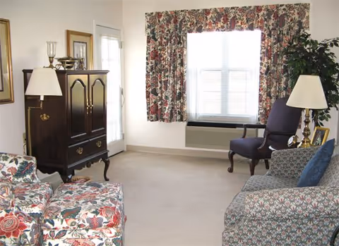 A living room with floral patterned armchairs and a sofa, a dark wooden cabinet, two table lamps, a window with floral curtains, and a potted plant in the corner.