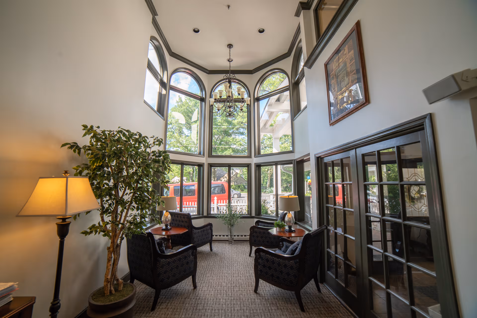 A bright and cozy sitting area with large arched windows letting in natural light. The room features four dark upholstered armchairs arranged around two small wooden tables with lamps. A tall potted plant and a chandelier hanging from the ceiling add to the inviting atmosphere. The walls are painted light with dark trim, and a framed picture hangs above a set of glass-paneled double doors.