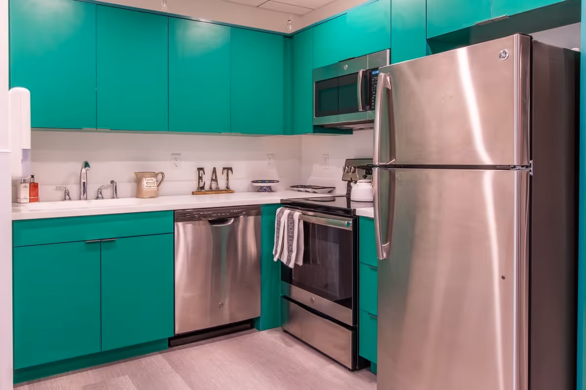 A modern kitchen with bright green cabinets, stainless steel appliances including a refrigerator, dishwasher, oven, and microwave. The countertop has a sink, a decorative pitcher, a bowl, and letters spelling 'EAT'.