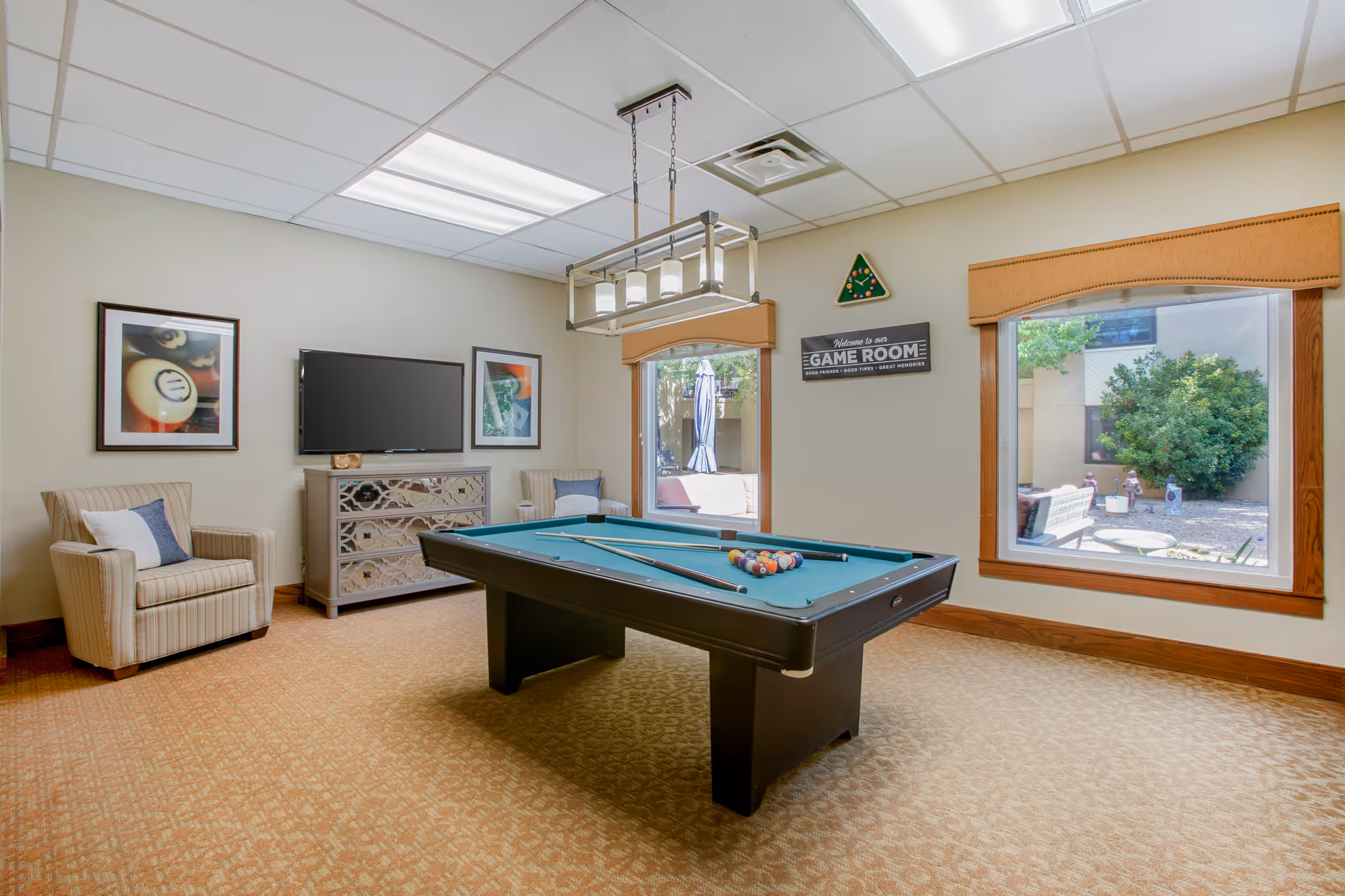 A well-lit game room with a pool table in the center, two pool cues resting on the table, and a rack of billiard balls arranged for a game. The room has beige walls, carpeted floor, two large windows with views of an outdoor garden area, a striped armchair with a pillow, a TV mounted on the wall above a decorative cabinet, and framed pictures on the walls. A sign on the wall reads 'Welcome to our GAME ROOM'.