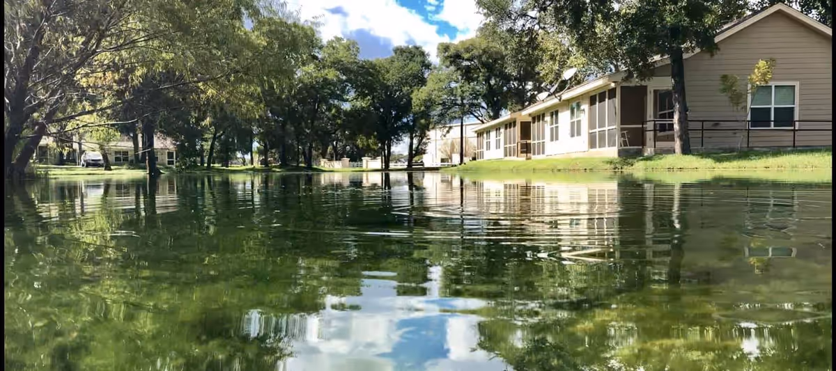 A serene outdoor scene at The Lodge at Leon Springs featuring a calm pond reflecting surrounding trees and buildings under a partly cloudy sky.