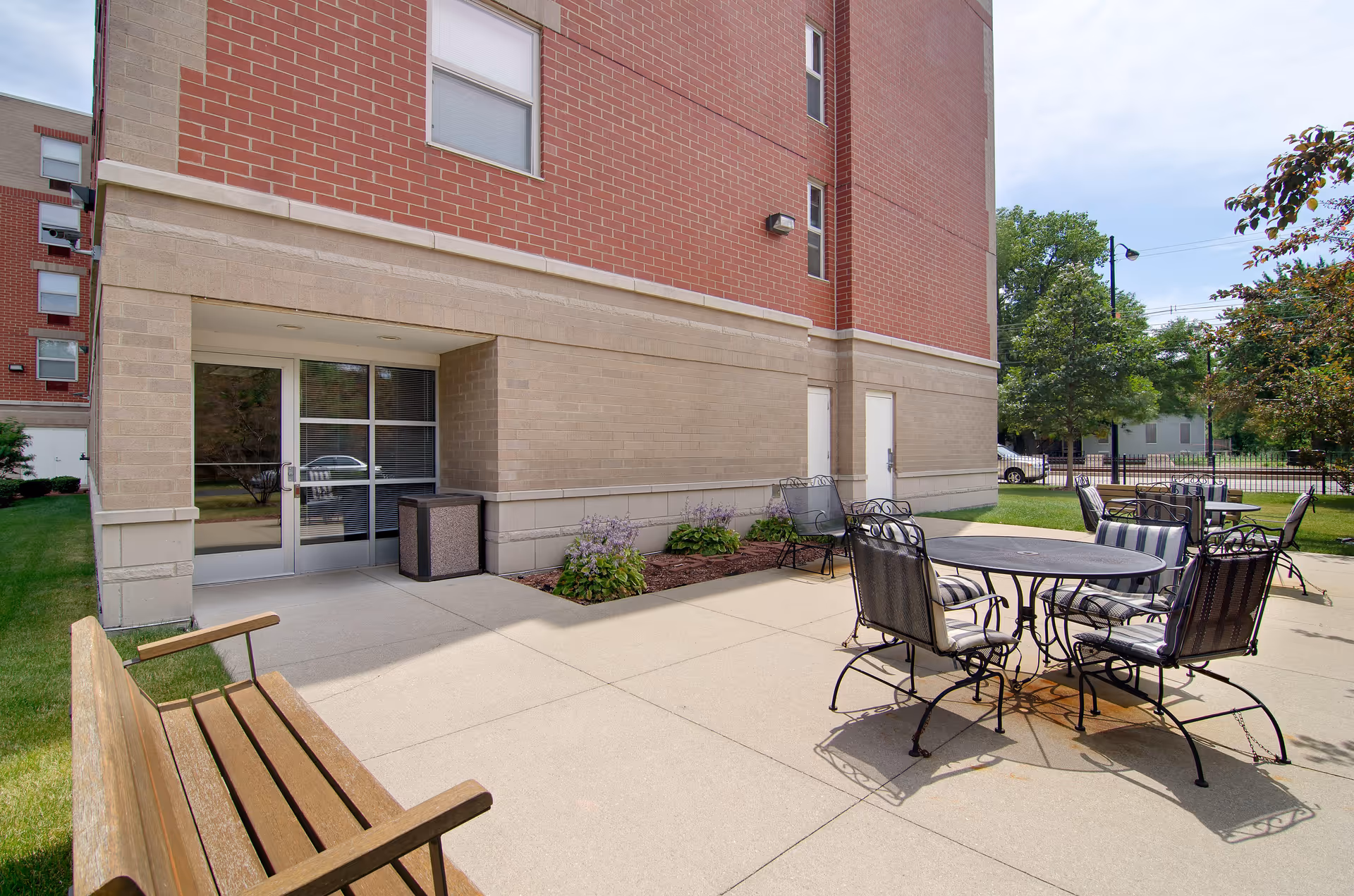 Outdoor patio area at Senior Suites of Rainbow Beach with metal tables and chairs featuring striped cushions, a wooden bench, and a building entrance with glass doors. The area is surrounded by grass, small plants, and trees under a partly cloudy sky.