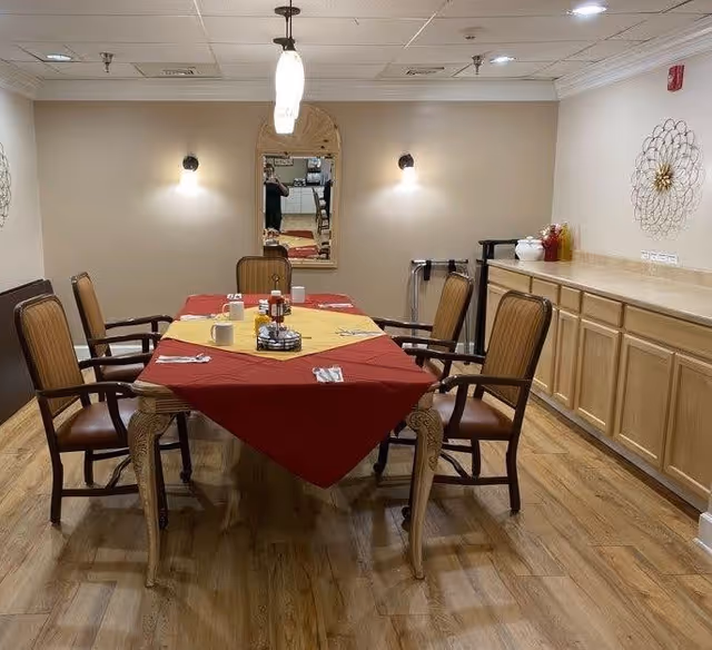 A dining room with a rectangular wooden table covered with a red and yellow tablecloth. Six wooden chairs with armrests surround the table. The table is set with cups, utensils, and condiments. The room has wooden flooring, beige walls, two wall-mounted lights, a decorative mirror, and a sideboard with decorative items on top.