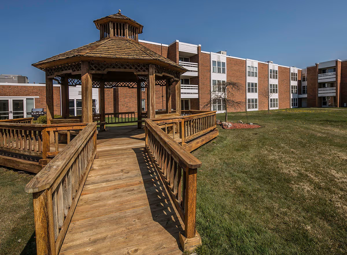 A wooden gazebo with a shingled roof and railings, situated on a grassy lawn in front of a multi-story brick building with white window frames and balconies under a clear blue sky.