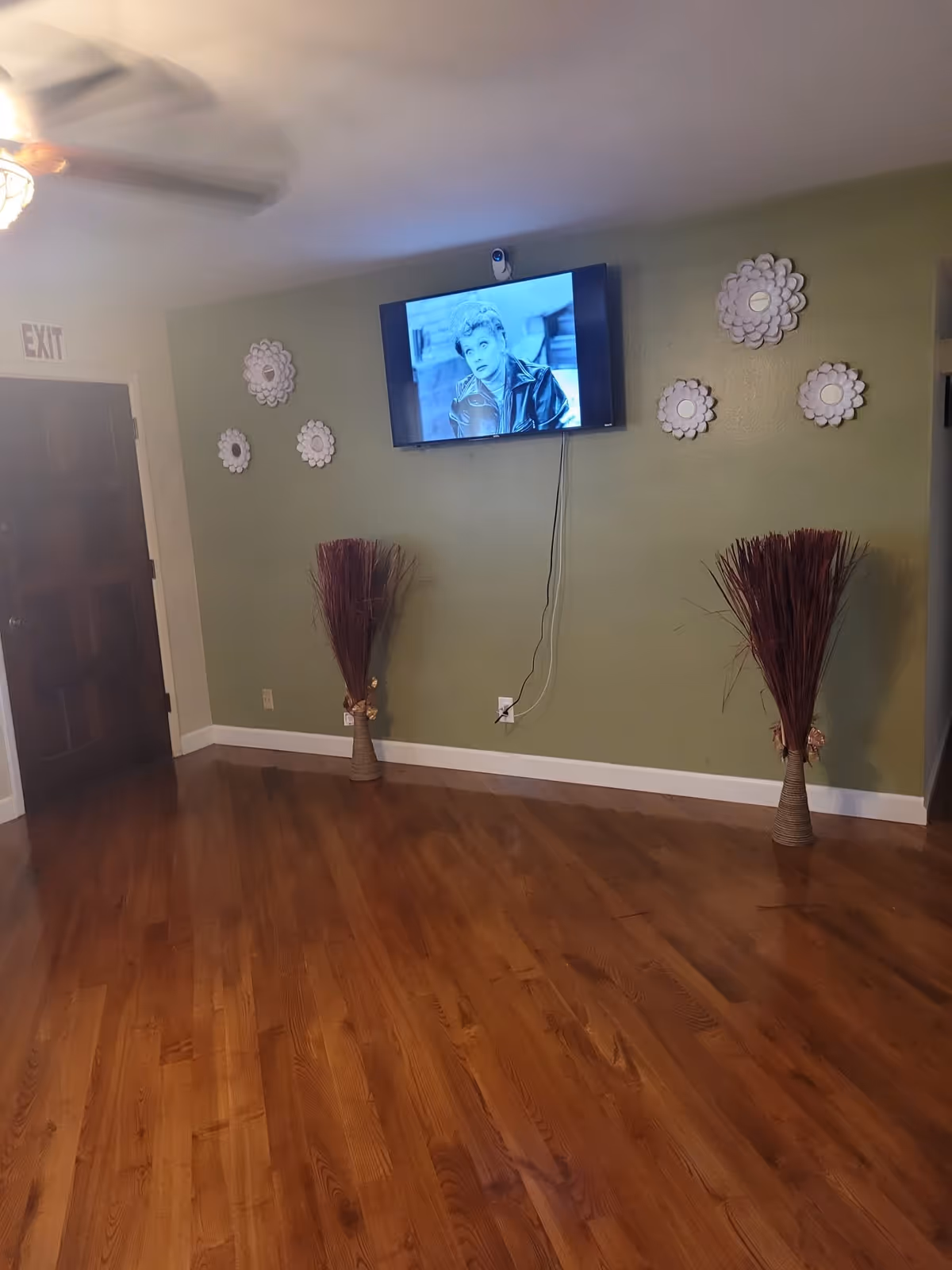 A room with polished wooden floors and light green walls. A flat-screen TV mounted on the wall displays a black and white image of a woman. Decorative white flower-shaped wall hangings are arranged around the TV. Two tall vases with dried branches are placed on the floor on either side of the TV. A dark wooden door with an exit sign above it is visible on the left side of the image. A ceiling fan with a light is partially visible at the top left corner.
