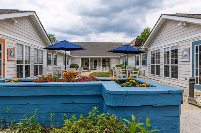 Courtyard of a memory care community with blue planter boxes, flowering beds, patio tables with blue umbrellas and surrounding single-story buildings.