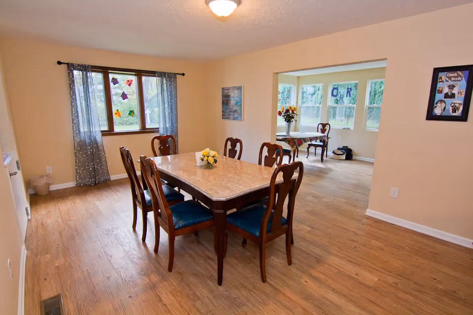 A dining room with a rectangular wooden table and six chairs with blue cushions. The room has light-colored walls and wood flooring. A window with blue patterned curtains is on the left wall, and there is an open doorway leading to a sunlit room with multiple windows, a small table, and chairs. The walls have framed pictures and decorations.