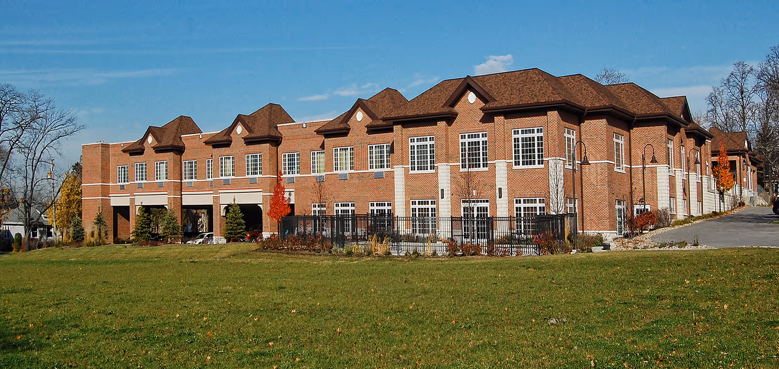 Exterior view of a large two-story brick building with multiple windows and a brown roof, surrounded by a grassy lawn and some trees with autumn foliage under a clear blue sky.