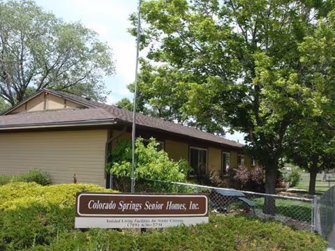 Exterior view of a single-story building surrounded by trees and bushes with a sign in front that reads 'Colorado Springs Senior Homes, Inc. Assisted Living Facilities for Senior Citizens (719) 636-2731'.