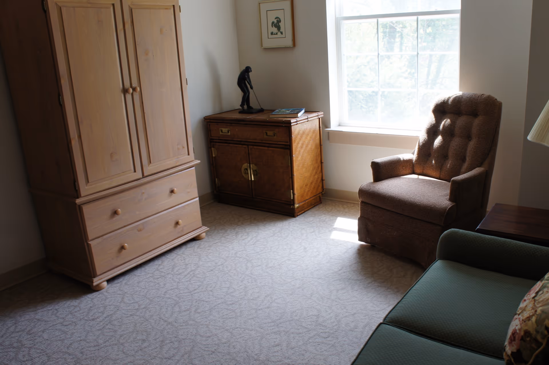 Sunny sitting area with a tufted armchair, wooden armoire, small cabinet and sofa by a window.