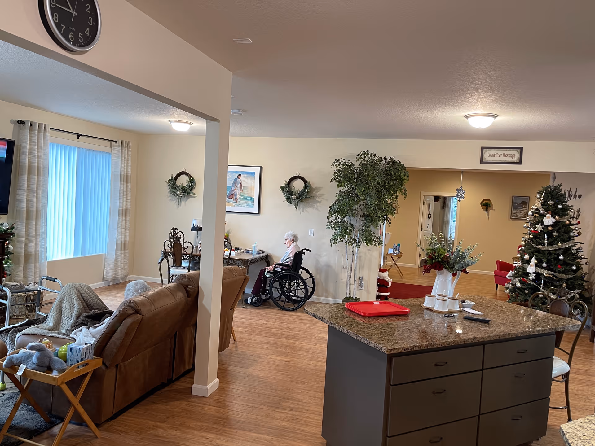 Interior view of a senior living facility showing a living room and dining area. There is a brown couch with a blanket and stuffed animal, a large clock on the wall, a person in a wheelchair near a table, a decorated Christmas tree, and a kitchen island with a red tray and some cups. The room has wooden flooring and light-colored walls with some framed pictures and wreaths.