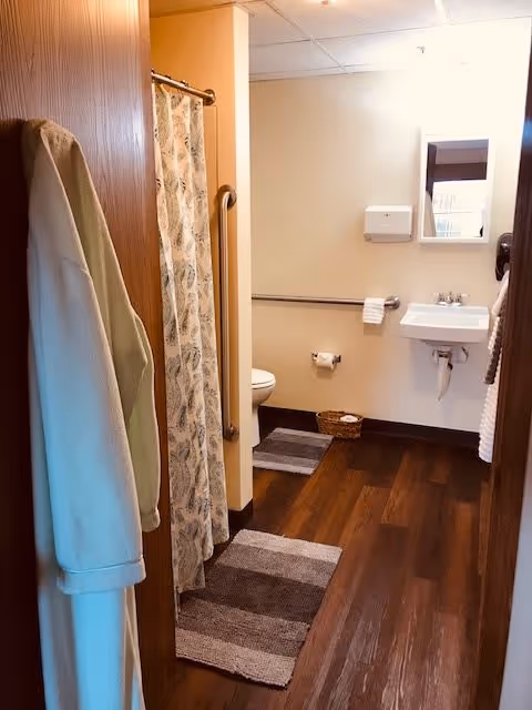 View into a bathroom with wooden flooring, a white robe hanging on the door, a shower with a patterned curtain, a toilet with grab bars, a white sink with a mirror above it, and a paper towel dispenser on the wall. There are two striped rugs on the floor and a small basket near the toilet.