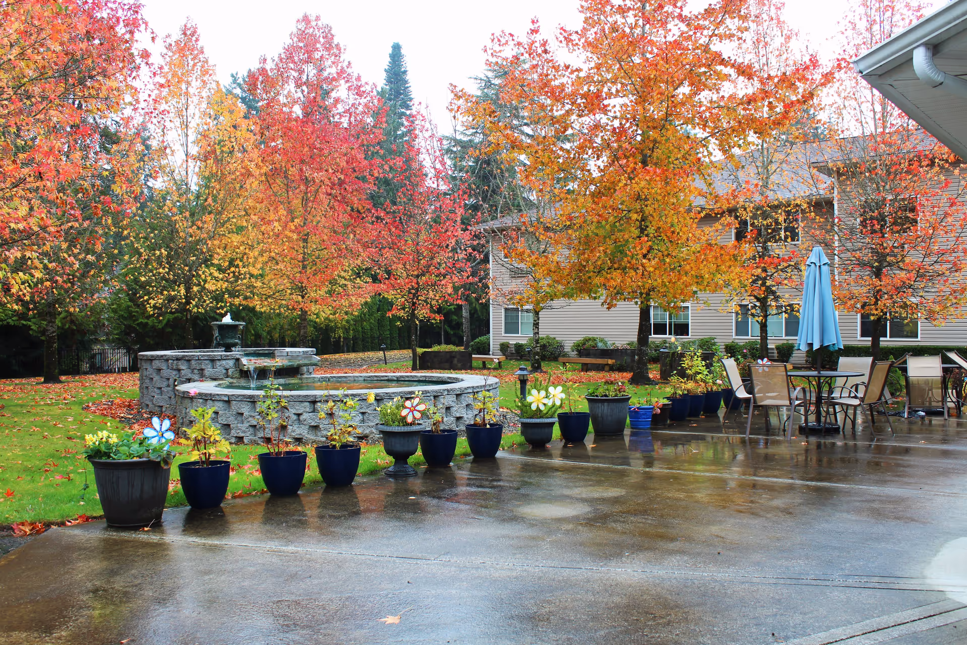 Outdoor courtyard with a stone fountain, potted plants and patio tables and chairs in front of a senior living building surrounded by autumn trees.