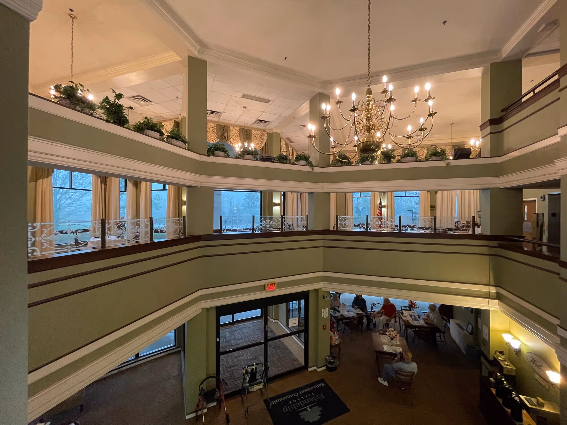Multi-level atrium interior with a large chandelier, wraparound balconies, potted plants, and seating areas with people at tables on the ground floor.