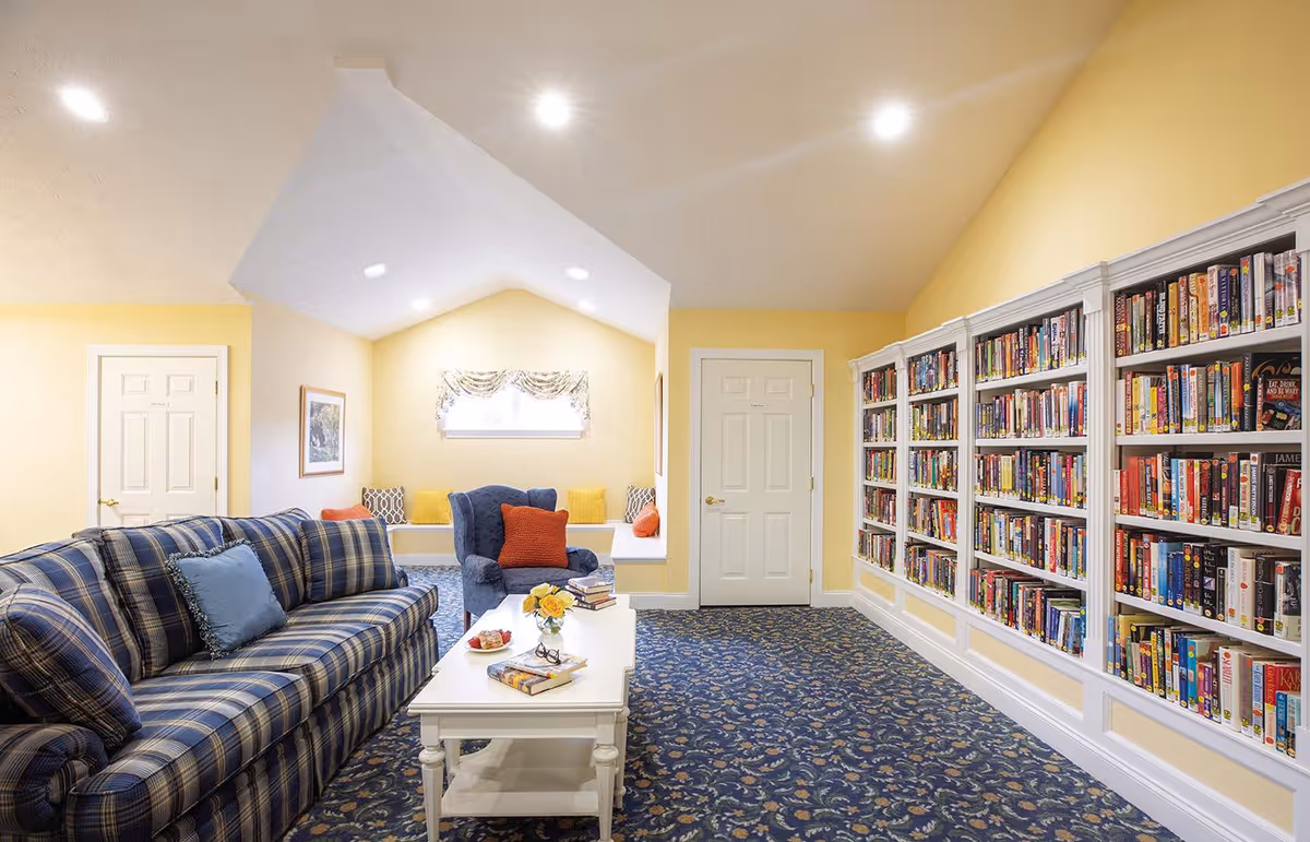 A cozy reading room with yellow walls and a blue patterned carpet. The room features a blue plaid sofa with a blue cushion, a blue armchair with an orange cushion, and a white coffee table with books and a small flower arrangement. Along one wall are built-in white bookshelves filled with books. There is a small window with a decorative valance above a built-in bench with several cushions. Two white doors are visible in the background.