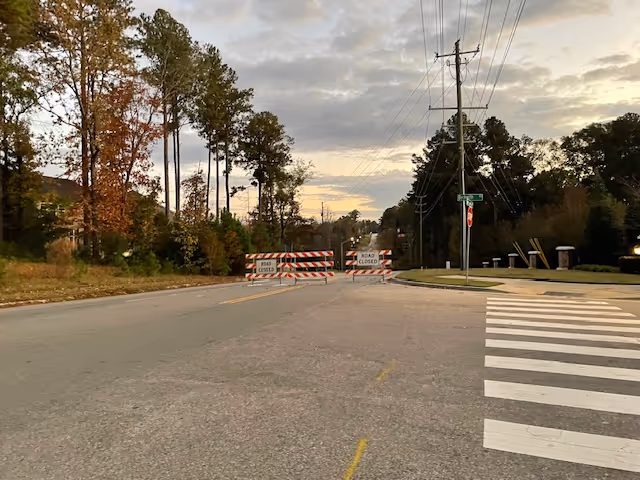 A road blocked by two 'Road Closed' barricades with trees and power lines on either side under a cloudy sky during sunset.