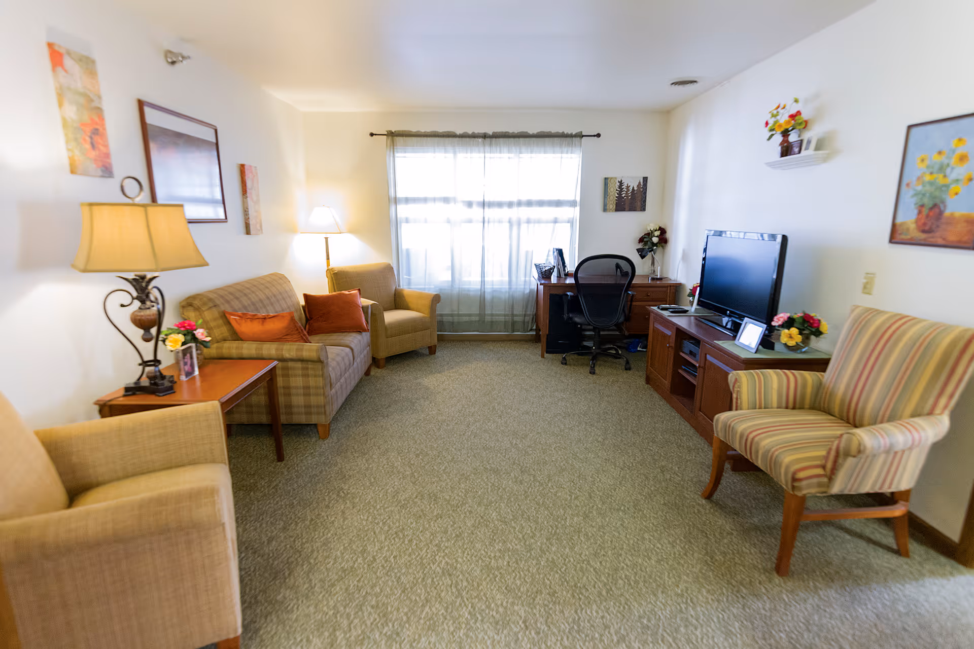 Bright common living room with sofas, armchairs, a TV on a media cabinet, and a desk by a window.