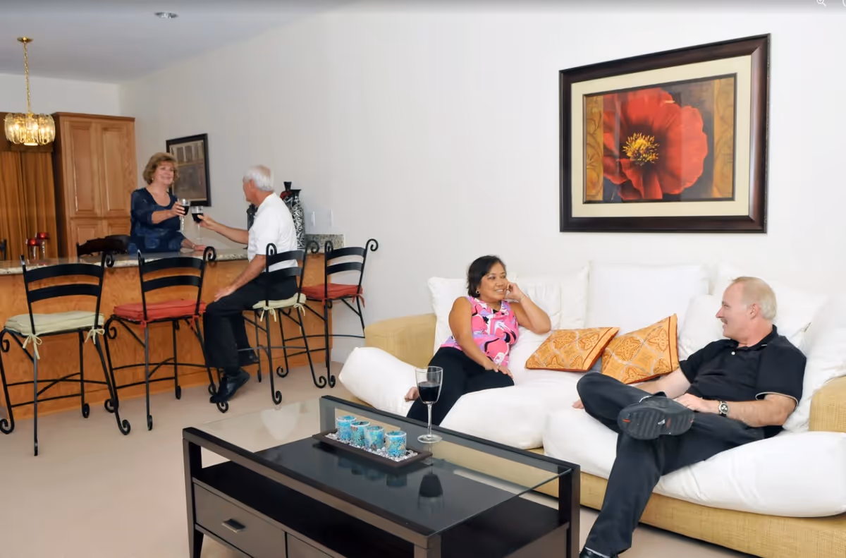 A living room and kitchen area in a retirement center where two women and two men are socializing. Two people are sitting on a white couch with orange patterned pillows, while the other two are seated on high chairs at a kitchen counter. The room features a large framed red flower painting on the wall and a black coffee table with decorative items.