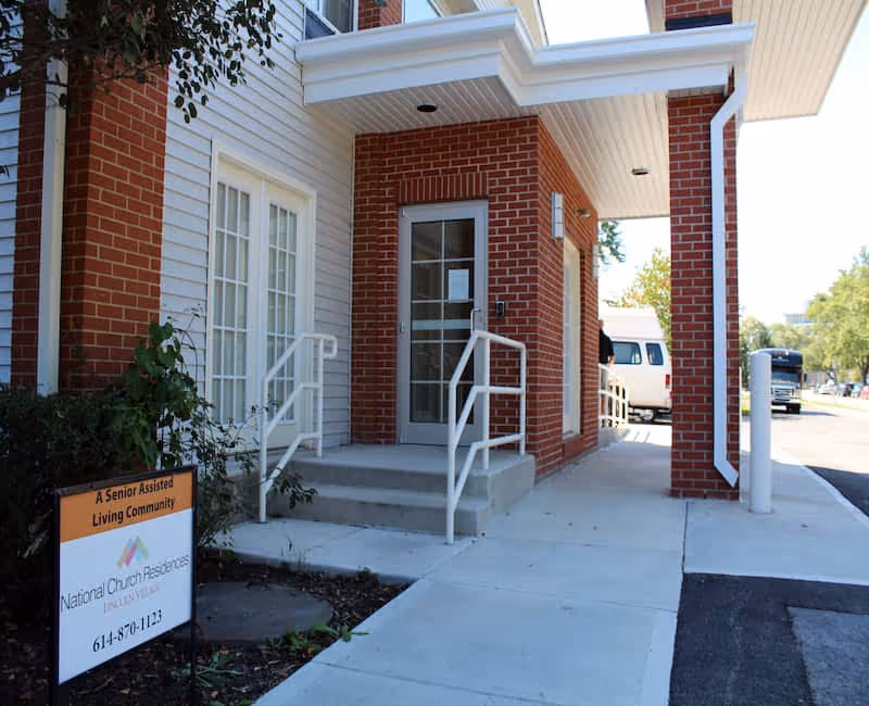 Front entrance of a brick-and-siding senior assisted living building with steps, handrails, and a small sign.