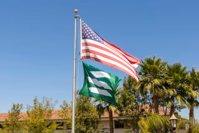 Two flags flying on a flagpole against a clear blue sky. The top flag is the United States flag, and below it is a green flag with a white design. In the background, there are palm trees, other greenery, and a building with a tiled roof.