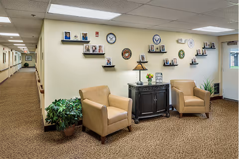 A cozy seating area in a senior living facility hallway with two tan armchairs, a small black cabinet with a lamp and flowers on top, and a wall decorated with framed photos and military insignia. The hallway extends to the left with handrails along the walls and a potted plant near the seating area.