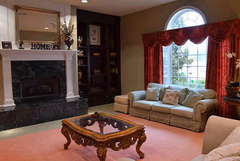 Cozy living room with sofa, ornate wooden coffee table, fireplace with mantle and built-in shelving beneath a large arched window.