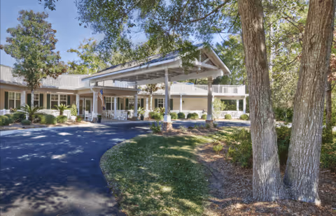 Exterior view of a senior living facility with a covered entrance driveway, surrounded by trees and landscaping. The building has a light-colored facade with white railings and an American flag near the entrance.