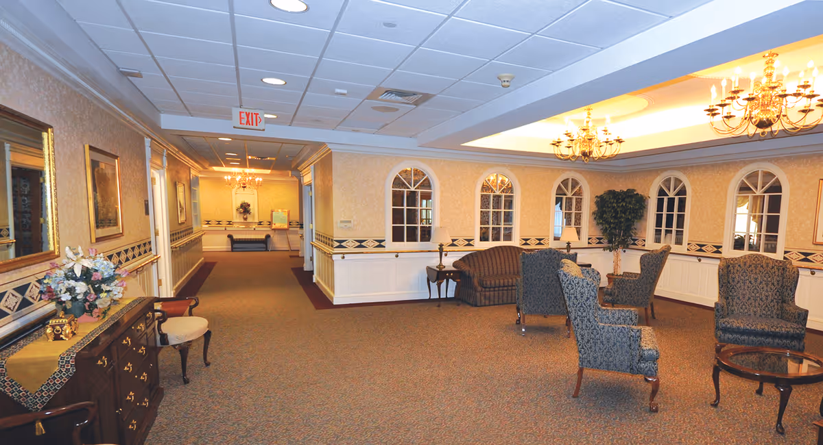 A spacious, well-lit interior common area with patterned carpet, several upholstered chairs and a sofa arranged around a glass-top coffee table. The walls are decorated with framed artwork and arched windows with mirrors. There are chandeliers hanging from the ceiling and a wooden sideboard with a floral arrangement on the left side of the image. A hallway extends into the background with an exit sign visible.