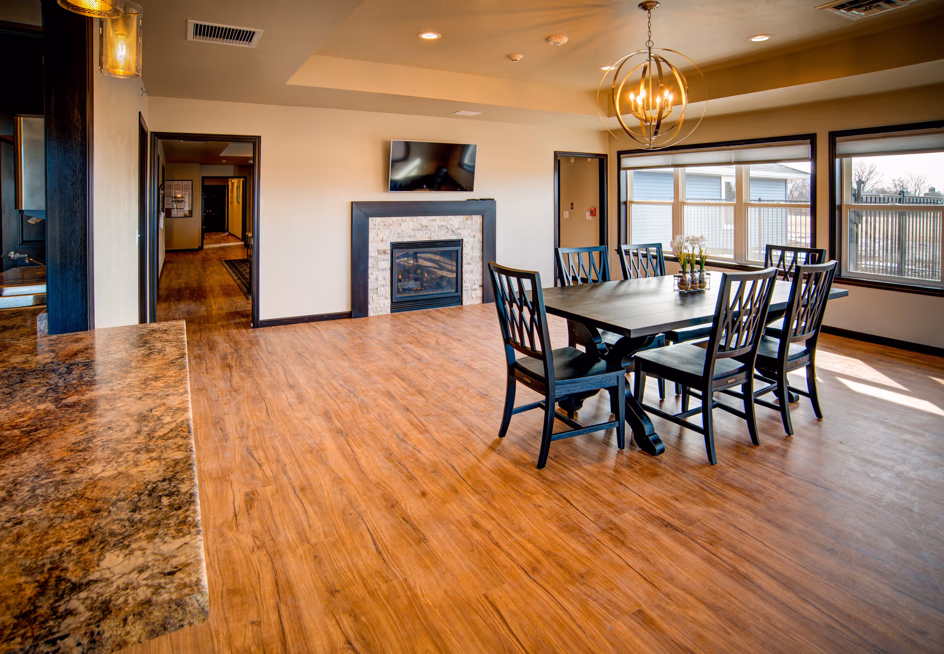 A bright dining room with a wooden table and six matching chairs. The room features large windows letting in natural light, a modern chandelier hanging above the table, a fireplace with a TV mounted above it, and wood flooring. There is a countertop visible on the left side and doorways leading to other rooms.