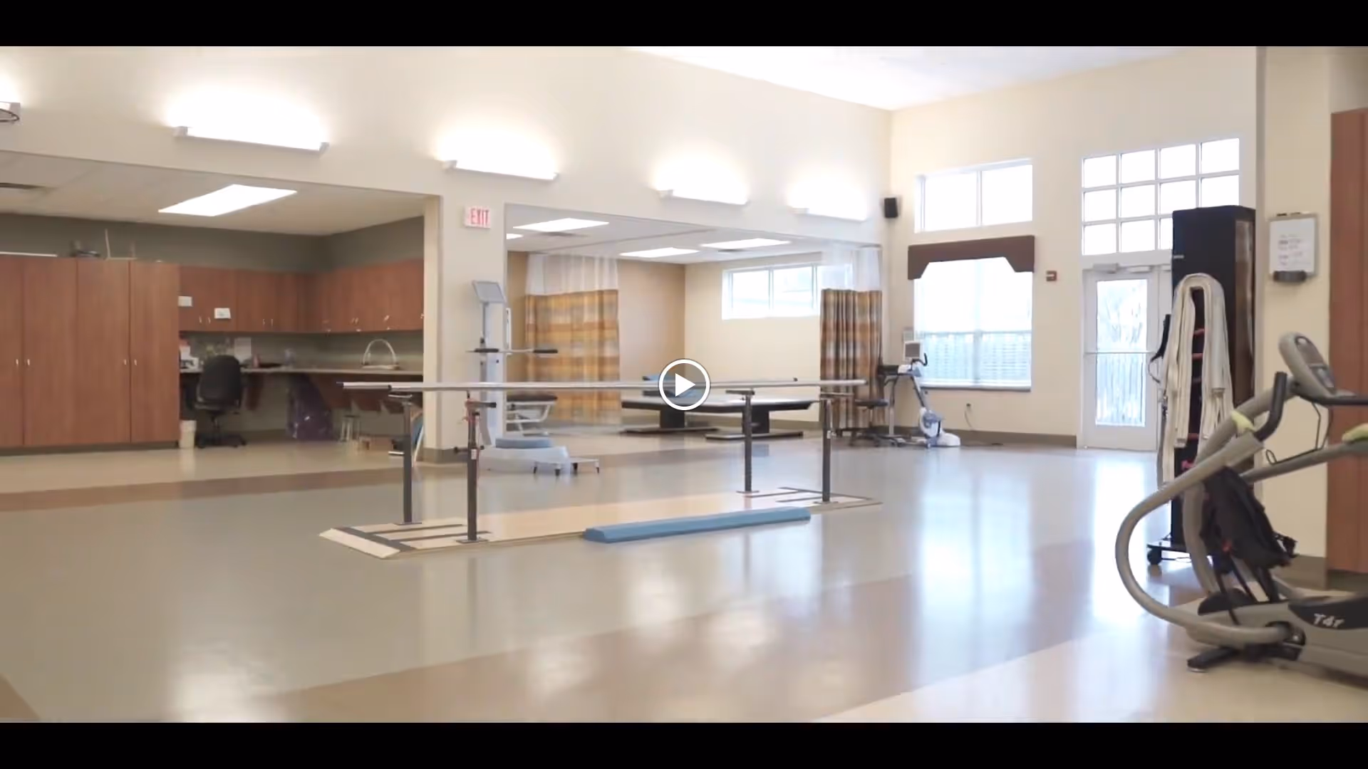 Bright rehabilitation/exercise room with parallel bars, exercise machines, cabinets, and windows in a senior living facility.