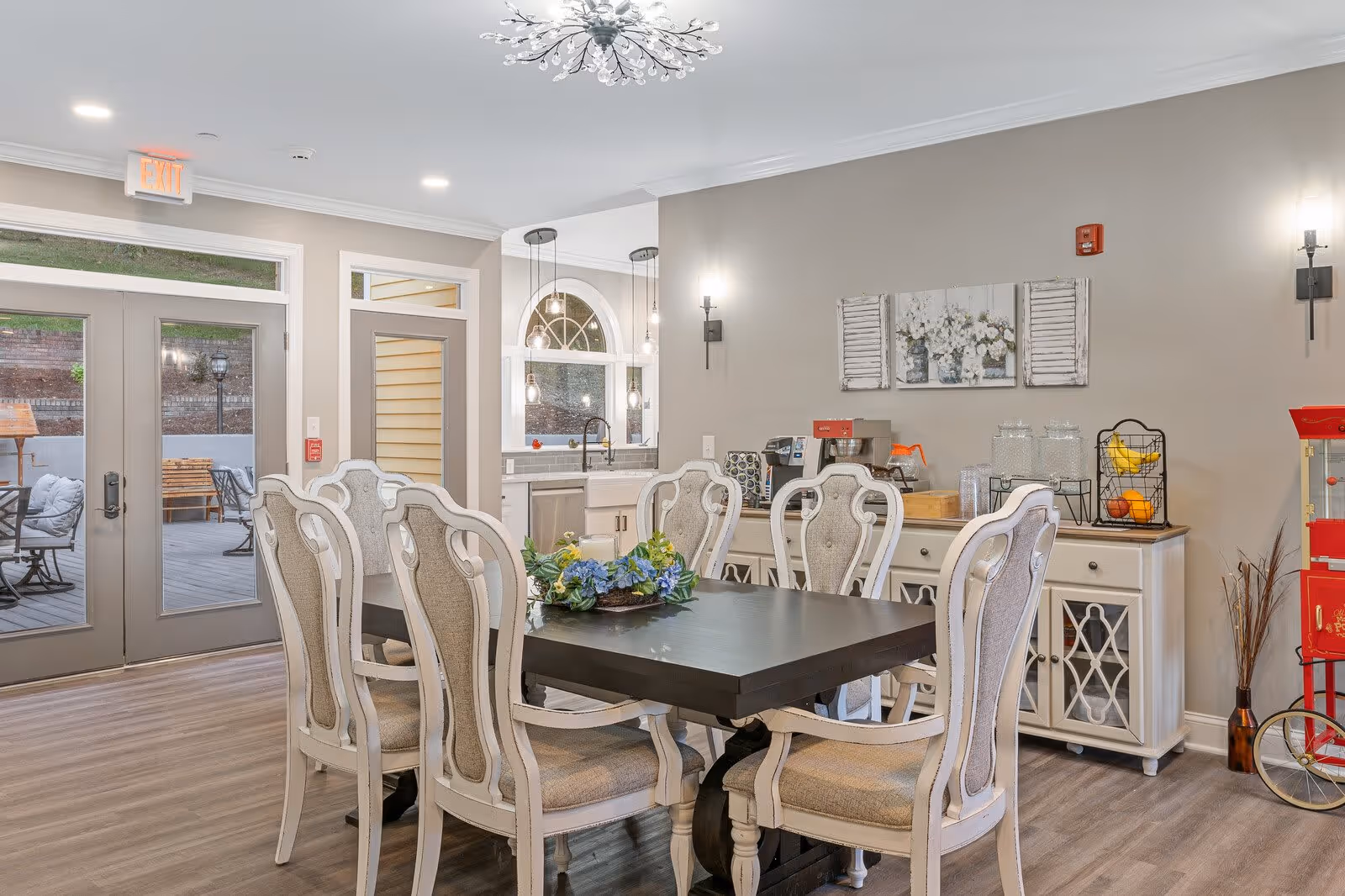 A bright dining room with a dark wooden table surrounded by eight white cushioned chairs. A floral centerpiece decorates the table. In the background, there is a white sideboard with a coffee maker, water dispensers, and fruit baskets. The room has light-colored walls, wood flooring, and a modern chandelier on the ceiling. Glass doors lead to an outdoor patio with seating visible outside.