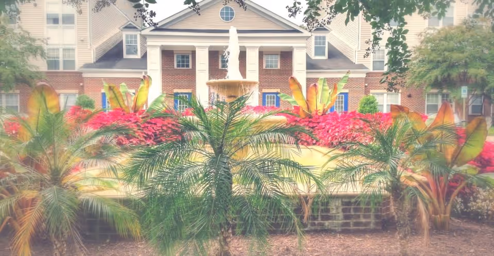 Front exterior of a senior living building featuring a columned entrance, central fountain, and colorful landscaped flowerbeds with palm plants.