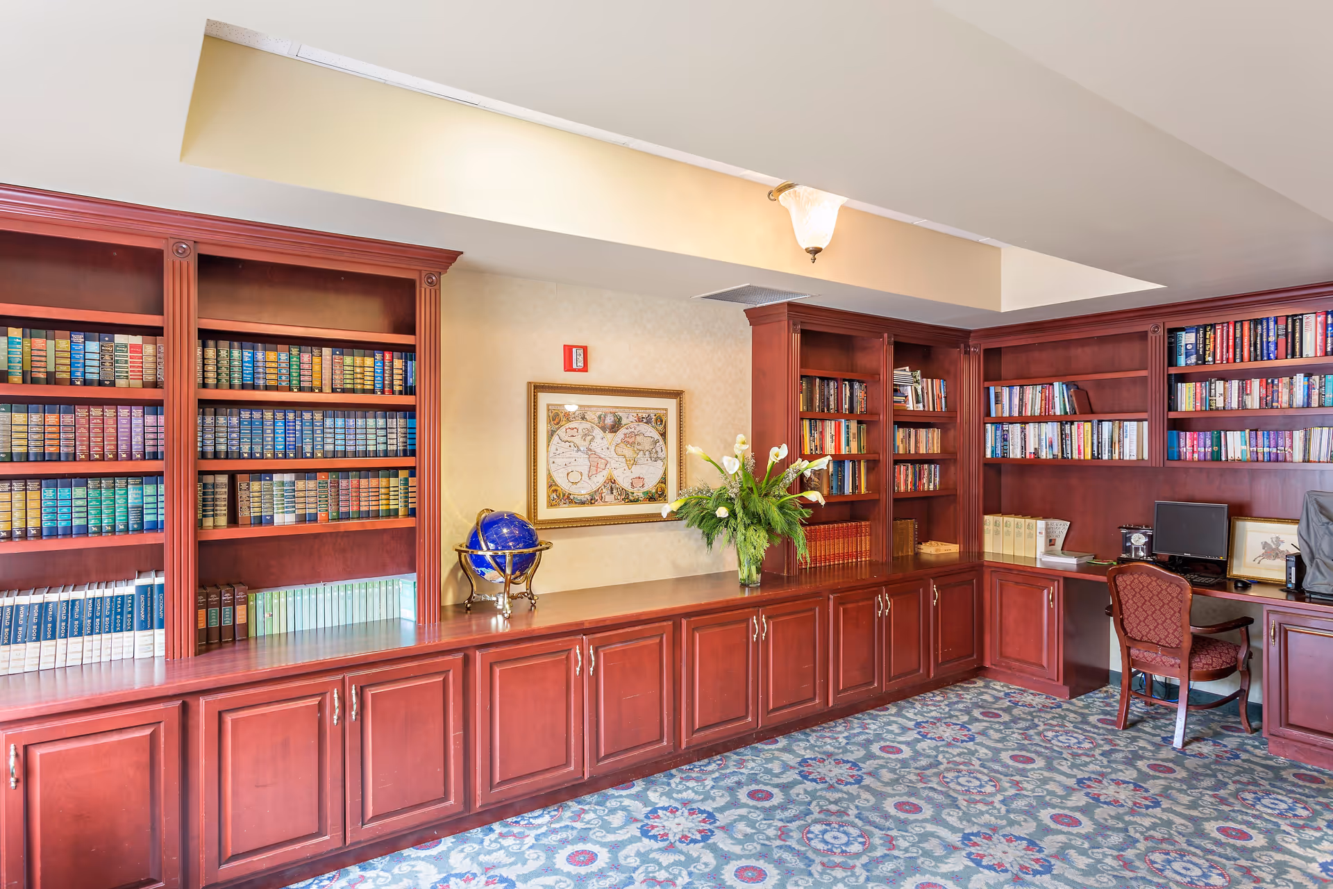 Interior view of a library or study room with wooden bookshelves filled with books, a globe on the counter, a framed world map on the wall, a vase with white flowers, a computer on a desk, and a patterned carpeted floor.