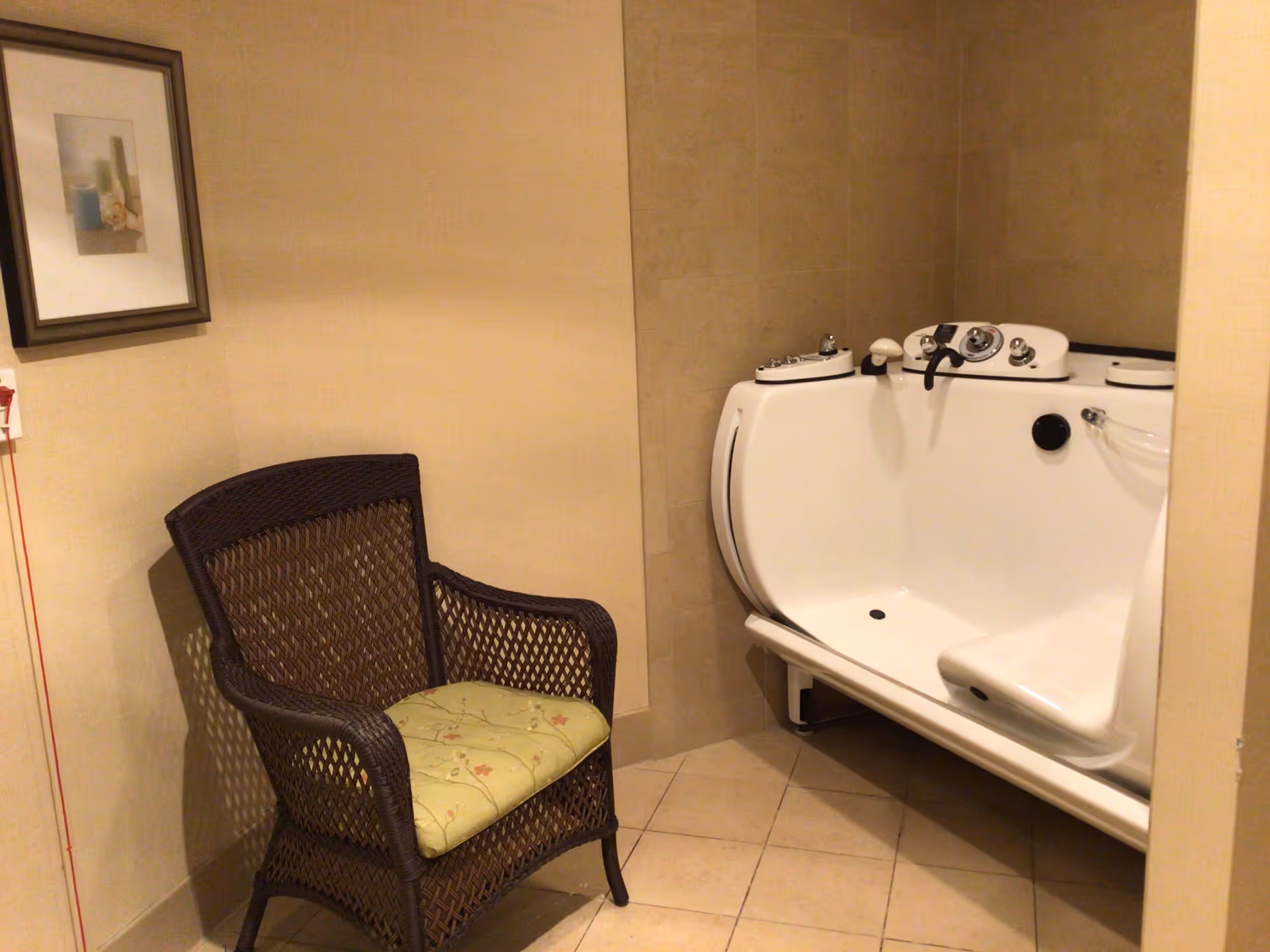 A small room with beige tiled walls and floor, featuring a white walk-in bathtub with multiple controls and a built-in seat. Next to the bathtub is a dark brown wicker chair with a light green cushion. A framed picture hangs on the wall above the chair.