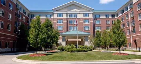 Brick senior living building with central entrance, lawn, and trees in front.