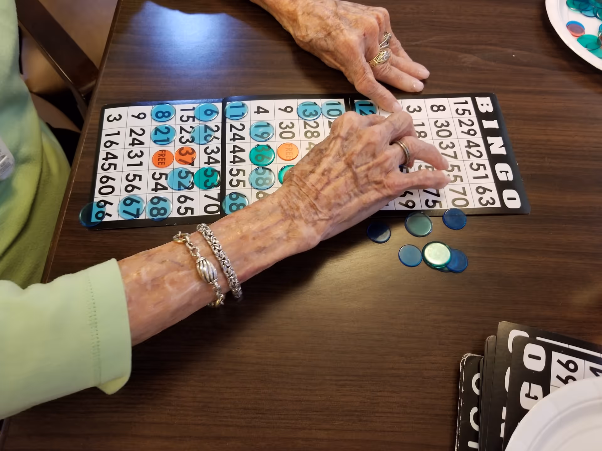 Two elderly hands placing translucent blue and green chips on a bingo card on a wooden table, with additional bingo cards and a paper plate nearby.