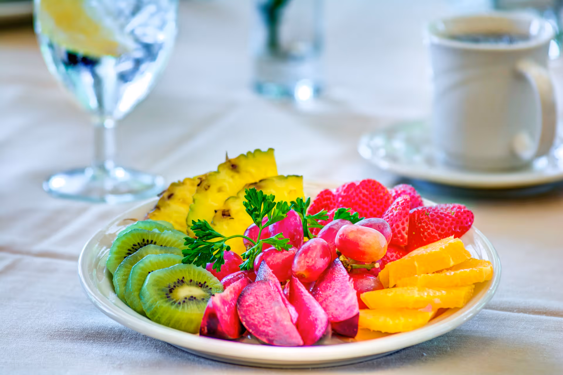 A plate of fresh fruit including sliced kiwi, pineapple, strawberries, grapes, orange slices, and plum wedges garnished with a sprig of parsley, placed on a table with a glass of water with lemon and a cup of coffee in the background.