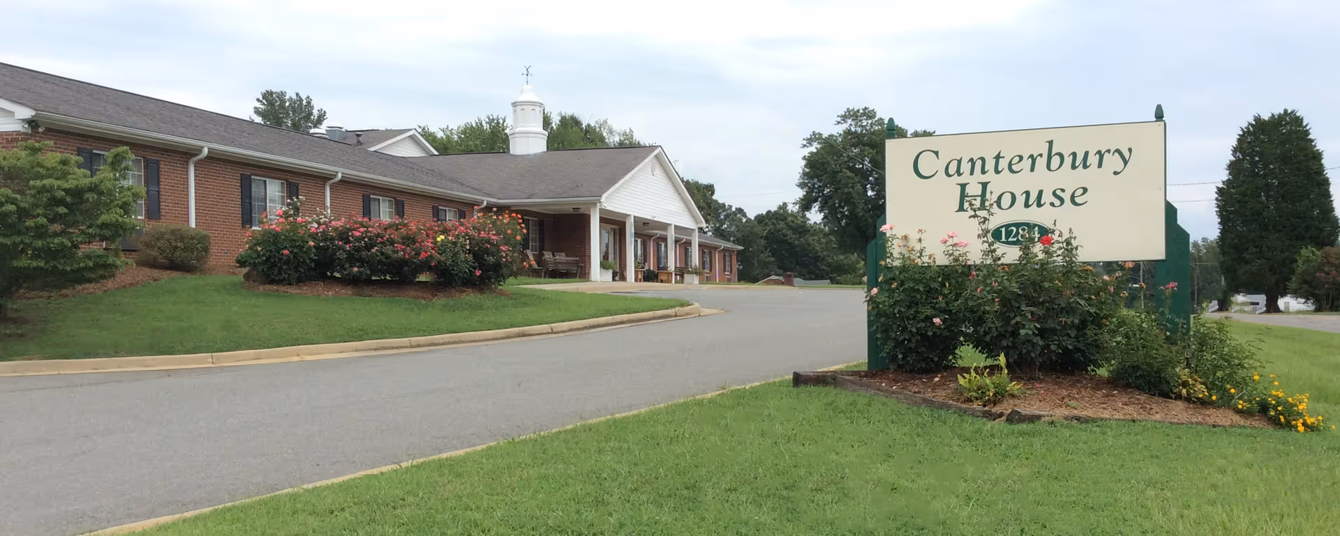 Exterior view of The Canterbury House facility showing a single-story brick building with a white entrance porch, surrounded by green lawns and bushes with flowers. A large sign with the name 'Canterbury House' and the number '1284' is visible near the driveway.