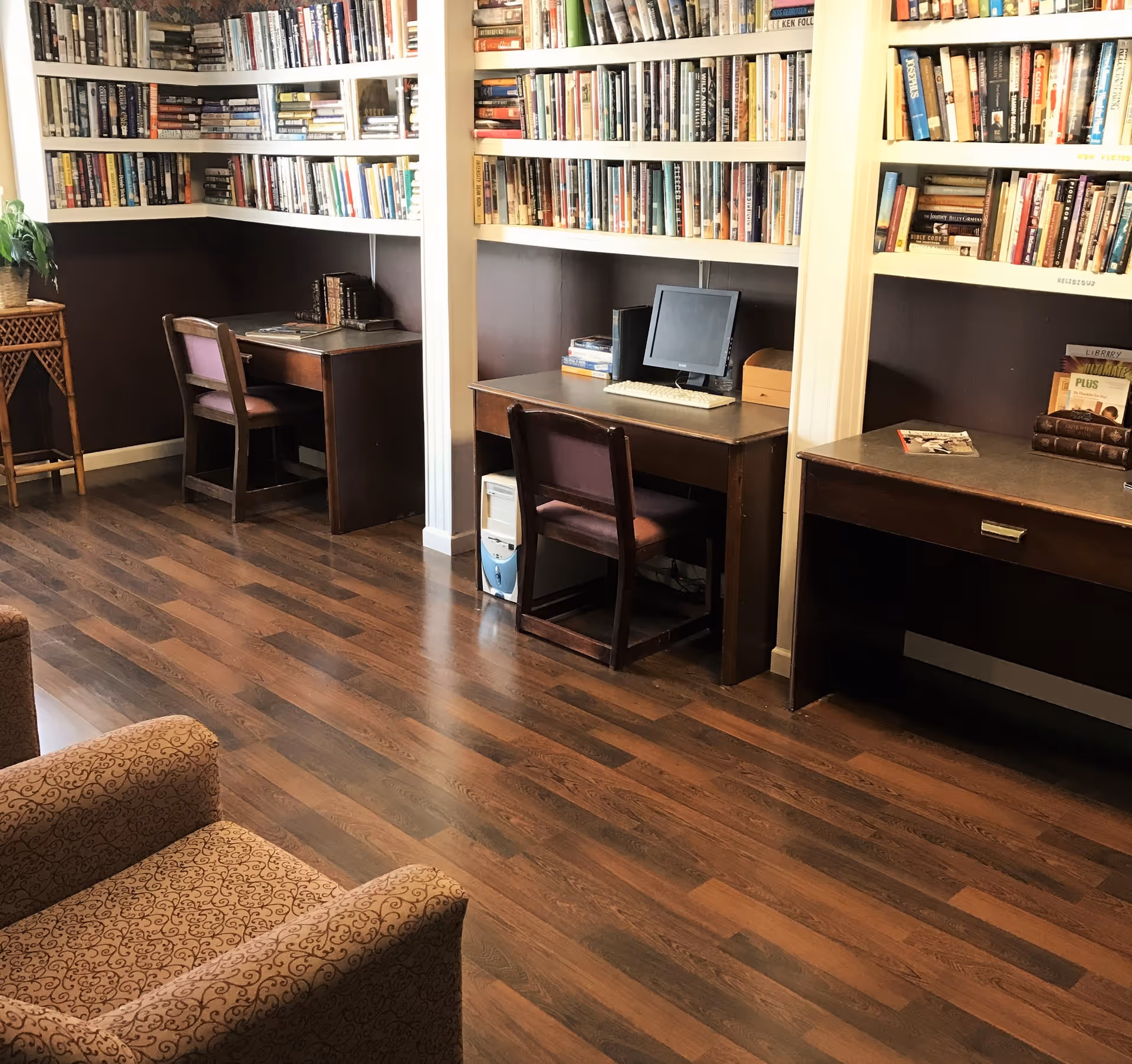 A cozy library or study area with wooden desks and chairs along the wall, filled with shelves of books. One desk has a computer with a monitor, keyboard, and tower. The floor is wooden, and there is a patterned armchair in the foreground.