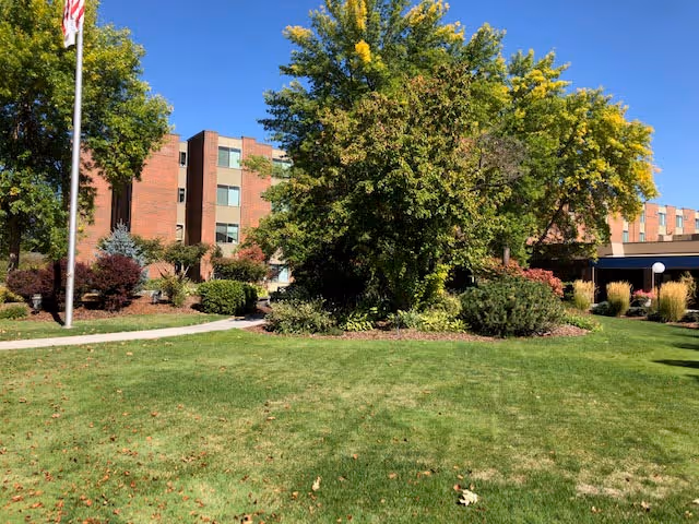 A well-maintained outdoor garden area with green grass, various bushes, and trees with some yellow flowers. In the background, there is a multi-story brick building under a clear blue sky. An American flag on a flagpole is visible on the left side.