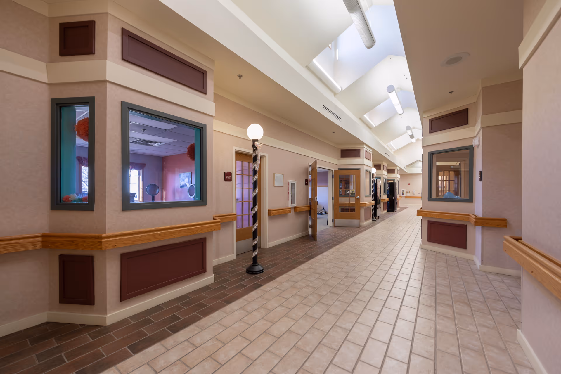 A wide, well-lit hallway in a senior living facility with tiled floors and beige walls accented with wooden trim. The hallway features several windows and doors leading to rooms, with some doors open. There are ceiling skylights and modern light fixtures along the corridor, and a decorative lamp post stands near the center.